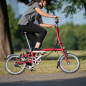 Preview: Eine Frau mit schwarzer Hose und grauem Shirt fährt rasant mit ihrem House Red Brompton Faltrad um die Kurve.