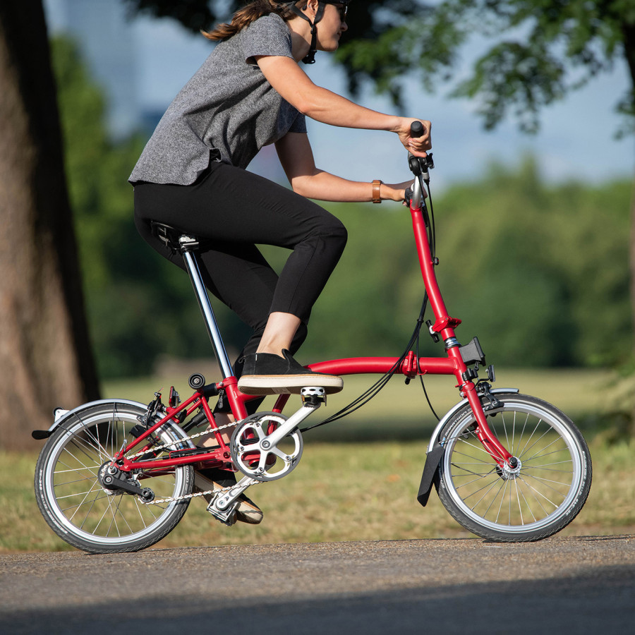 Eine Frau mit schwarzer Hose und grauem Shirt fährt rasant mit ihrem House Red Brompton Faltrad um die Kurve.