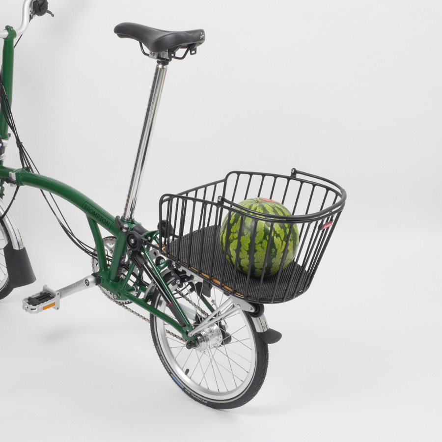 Der Rixen & Kaul Gepäckträgerkorb KLICKfix Alumino GT in Black ist auf den Gepäckträger eines grünen Brompton Faltrades geklippt, im Korb liegt eine Melone, darunter ein schwarzer Schaumstoffschutz