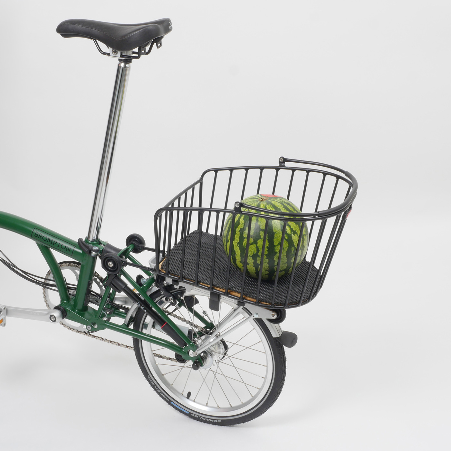 Der Rixen & Kaul Gepäckträgerkorb KLICKfix Alumino GT in Black ist auf den Gepäckträger eines grünen Brompton Faltrades geklippt, im Korb liegt eine Melone