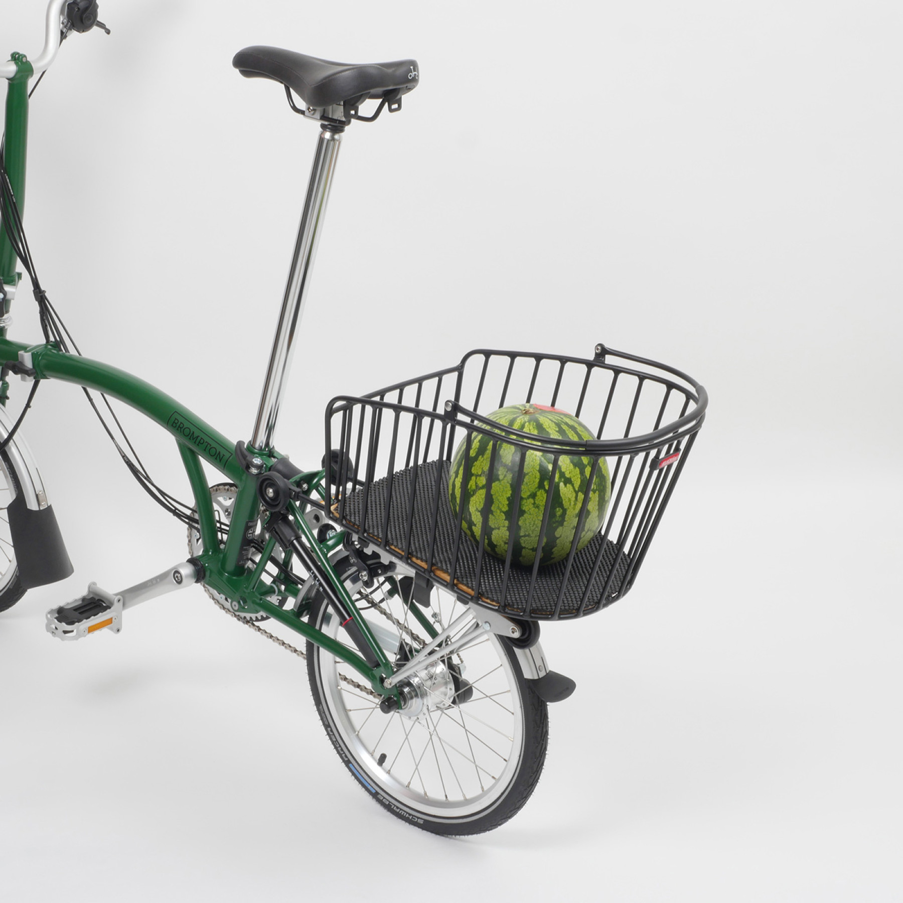 Der Rixen & Kaul Gepäckträgerkorb KLICKfix Alumino GT in Black ist auf den Gepäckträger eines grünen Brompton Faltrades geklippt, im Korb liegt eine Melone, darunter ein schwarzer Schaumstoffschutz